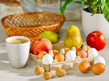 Açık karton bir kutuda kahverengi ve beyaz tavuk yumurtaları. Sabah güneşinde kahvaltı hazırlıyorum. Masada kahve, meyve ve çiçek var. Paskalya