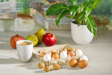 Açık karton bir kutuda kahverengi ve beyaz tavuk yumurtaları. Sabah güneşinde kahvaltı hazırlıyorum. Masada kahve, meyve ve çiçek var. Paskalya