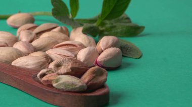 Yeşil yapraklar ve kahverengi tahta kaşıkla süslenmiş Pistachios veya Pista fındıkları. Yeşil arka plan, üst manzara. Düz yatıyordu.