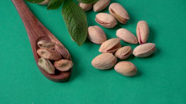 Yeşil yapraklar ve kahverengi tahta kaşıkla süslenmiş Pistachios veya Pista fındıkları. Yeşil arka plan, üst manzara. Düz yatıyordu.