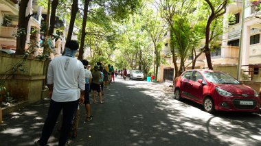 INDIA - 08 Mayıs 2020: Yeşil ağaçların gölgesinde park etmiş arabalarla eski caddenin yanından geçen kalabalık  