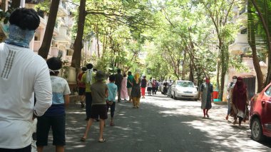 INDIA - 08 Mayıs 2020: Yeşil ağaçların gölgesinde park etmiş arabalarla eski caddenin yanından geçen kalabalık  