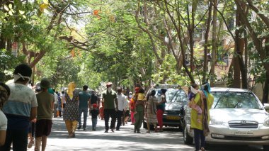 INDIA - 08 Mayıs 2020: Yeşil ağaçların gölgesinde park etmiş arabalarla eski caddenin yanından geçen kalabalık  