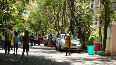 INDIA - 08 Mayıs 2020: Yeşil ağaçların gölgesinde park etmiş arabalarla eski caddenin yanından geçen kalabalık  