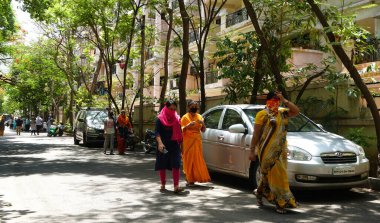 INDIA - 08 Mayıs 2020: Yeşil ağaçların gölgesinde park etmiş arabalarla eski caddenin yanından geçen kalabalık  