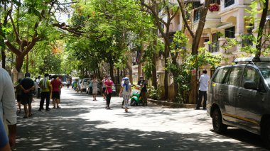 INDIA - 08 Mayıs 2020: Yeşil ağaçların gölgesinde park etmiş arabalarla eski caddenin yanından geçen kalabalık  