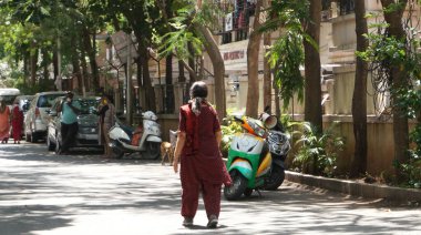 INDIA - 08 Mayıs 2020: Yeşil ağaçların gölgesinde park etmiş arabalarla eski caddenin yanından geçen kalabalık  