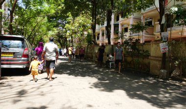 INDIA - 08 Mayıs 2020: Yeşil ağaçların gölgesinde park etmiş arabalarla eski caddenin yanından geçen kalabalık  