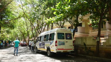 INDIA - 08 Mayıs 2020: Yeşil ağaçların gölgesinde park etmiş arabalarla eski caddenin yanından geçen kalabalık  