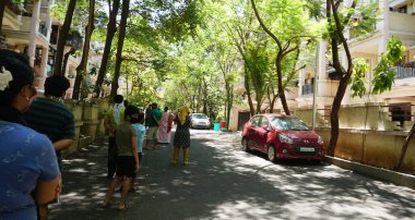 INDIA - 08 Mayıs 2020: Yeşil ağaçların gölgesinde park etmiş arabalarla eski caddenin yanından geçen kalabalık  