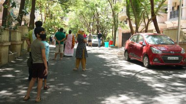 INDIA - 08 Mayıs 2020: Yeşil ağaçların gölgesinde park etmiş arabalarla eski caddenin yanından geçen kalabalık  