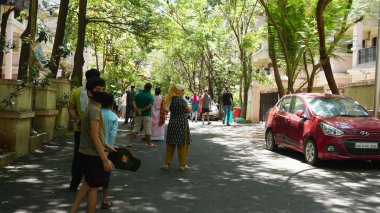 INDIA - 08 Mayıs 2020: Yeşil ağaçların gölgesinde park etmiş arabalarla eski caddenin yanından geçen kalabalık  