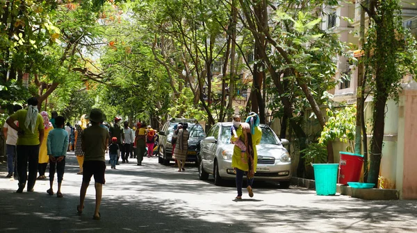 INDIA - 08 Mayıs 2020: Yeşil ağaçların gölgesinde park etmiş arabalarla eski caddenin yanından geçen kalabalık  