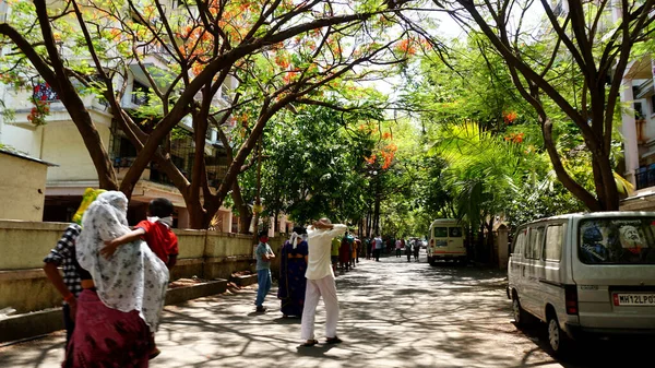 INDIA - 08 Mayıs 2020: Yeşil ağaçların gölgesinde park etmiş arabalarla eski caddenin yanından geçen kalabalık  