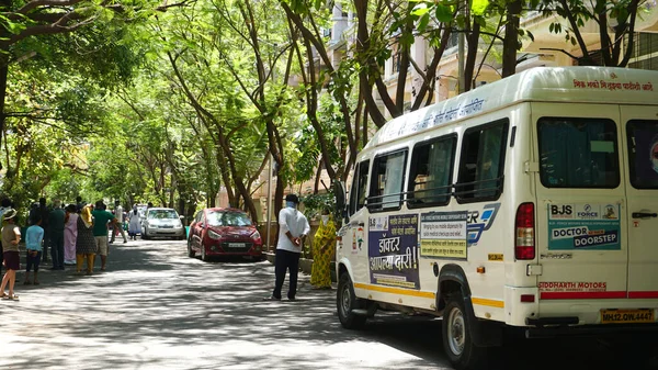 INDIA - 08 Mayıs 2020: Yeşil ağaçların gölgesinde park etmiş arabalarla eski caddenin yanından geçen kalabalık  