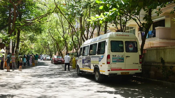 INDIA - 08 Mayıs 2020: Yeşil ağaçların gölgesinde park etmiş arabalarla eski caddenin yanından geçen kalabalık  