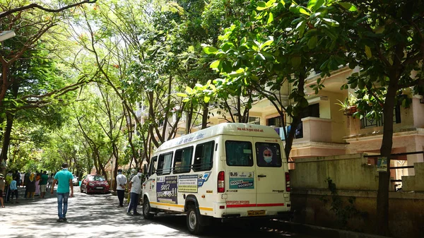 INDIA - 08 Mayıs 2020: Yeşil ağaçların gölgesinde park etmiş arabalarla eski caddenin yanından geçen kalabalık  