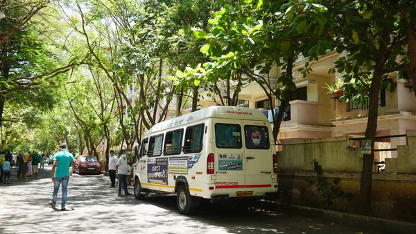 INDIA - 08 Mayıs 2020: Yeşil ağaçların gölgesinde park etmiş arabalarla eski caddenin yanından geçen kalabalık  