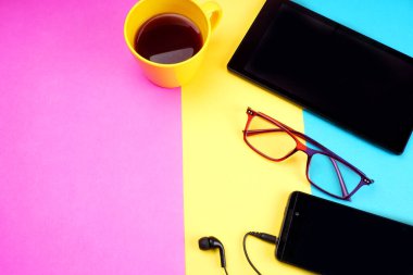Digital tablet, coffee cup, glasses and smartphone with headphones on colorful background
