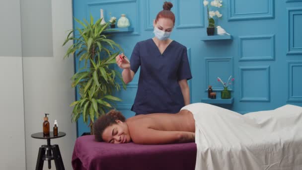 Masérka v lékařské masce obličeje nalévání oleje na klienta žena zpět dovnitř — Stock video