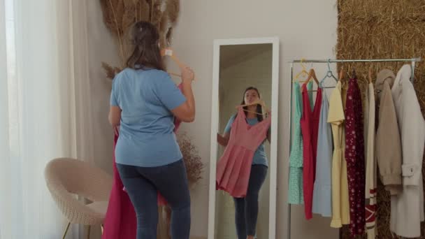 Plus-size kvinde sammenligner kjoler stående foran spejlet indendørs – Stock-video