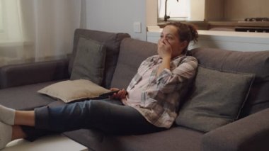 Yetişkin bir kadın uzaktan kumandayı tutuyor, televizyon izliyor, evde esniyor.