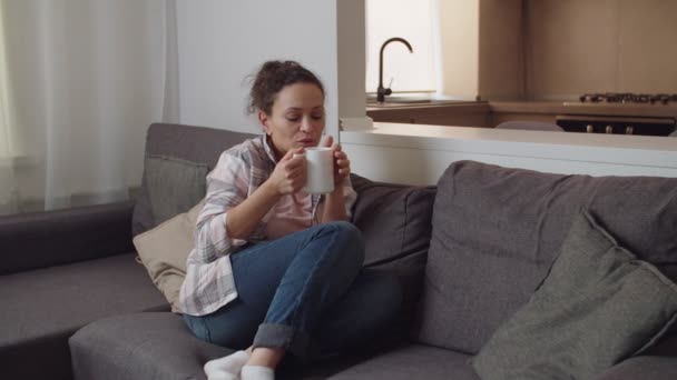 Mulher segurando caneca branca bebendo bebida quente sentado no sofá em casa — Vídeo de Stock