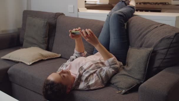 Mulher adulta tomando selfie usando aplicativo móvel enquanto deitada no sofá interior — Vídeo de Stock