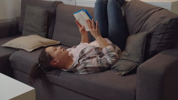 Доросла жінка читає книгу, кидаючи ноги над диваном в приміщенні — стокове відео