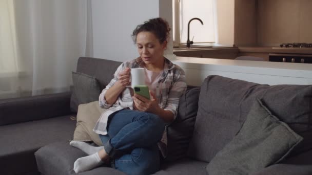 Mulher usando smartphone, bebendo bebida quente com caneca branca dentro de casa — Vídeo de Stock