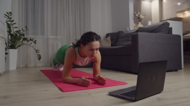 Mujer haciendo ejercicio de tablón, viendo video tutorial en el portátil en el interior — Vídeo de stock