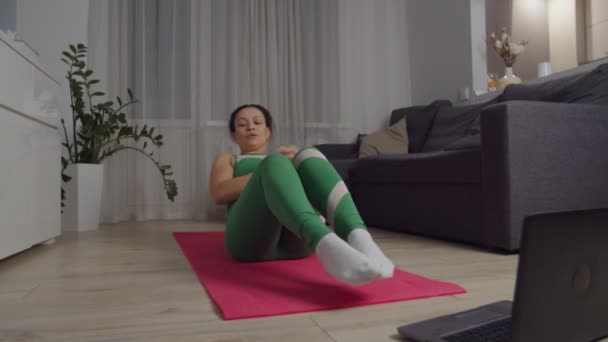 Mulher realizando exercícios abdominais sentado no tapete de ioga, usando laptop indoor — Vídeo de Stock