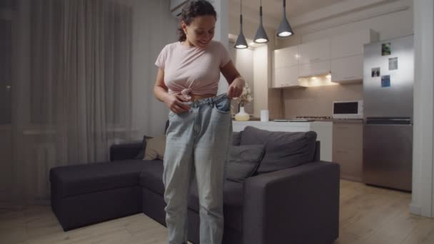 Mujer usando pantalones vaqueros sueltos, admirando el cuerpo después de la pérdida de peso en interiores — Vídeos de Stock