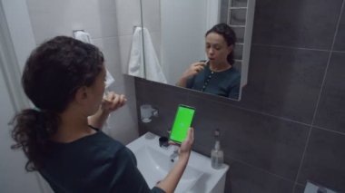 Siyah kadın dişlerini fırçalarken krom anahtarlı akıllı telefona bakıyor.