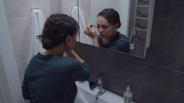 Woman applying mascara standing in front of vanity mirror indoors — Stockvideo