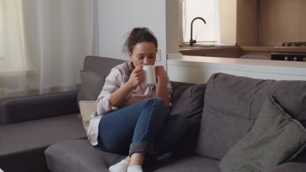 Adulto feminino relaxante com caneca quente de alguma bebida no sofá dentro de casa — Vídeo de Stock