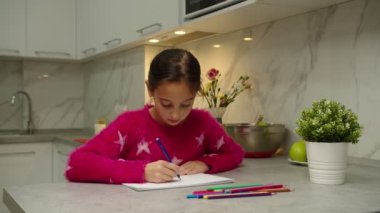 Evdeki eskiz defterine pembe kazaklı sevimli bir kız resim çiziyor.