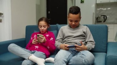 Çok ırklı çocuklar akıllı telefon kullanıyor, evdeki kanepede oturuyor..