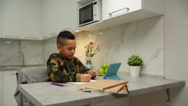 Симпатичный мальчик, пишущий в тетрадке, используя цифровой планшет во время домашнего обучения — стоковое видео