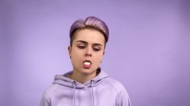 Mor kapüşonlu, kısa menekşe saç stili sakızlı, ilgisiz görünüşlü, tek renkli arka planda sakız şişiren çağdaş kadın. Yavaş çekim