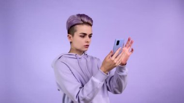 Mor kapüşonlu, kısa menekşe saç stili, akıllı telefona bakan, internet sitesinde şok edici haberler okuyan, menekşe arkaplanından izole edilmiş başını kavrayan, konsantre olmuş genç bir kadının stüdyo fotoğrafı.
