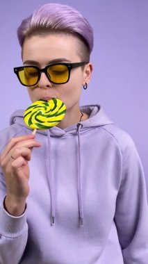 Mor kumaşlı, kısa menekşe saçlı, lolipop yalayan, tek renkli arka planda zevkle şeker yiyen genç kadın neslinin dikey görüntüsü.