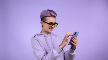 Kısa boyalı mor saçlı, menekşe rengi kazaklı, sarı güneş gözlüklü neşeli, genç bir kadın. Komik notlar bulduğuna şaşırmış, gülüyor, tek renkli arka planda izole akıllı telefon kullanıyor.