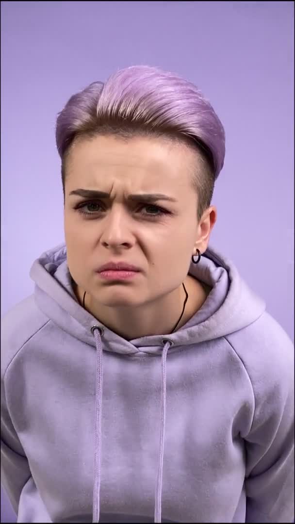一个头戴紫色帽衫的多愁善感的年轻女性 表现出厌恶的表情 不满和恐惧 由于对室内孤立的单色背景的厌恶反应而做着厌恶的表情 — 图库视频影像
