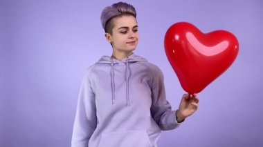 Moda tarzı kısa mor saç stiline sahip pozitif Igbt insanı kalp şeklinde kırmızı balon, aşk sembolü gösteriyor, kameraya mutlu bir şekilde bakıyor, kapalı bir menekşe arka planda kapüşonlu bir şey giyiyor.