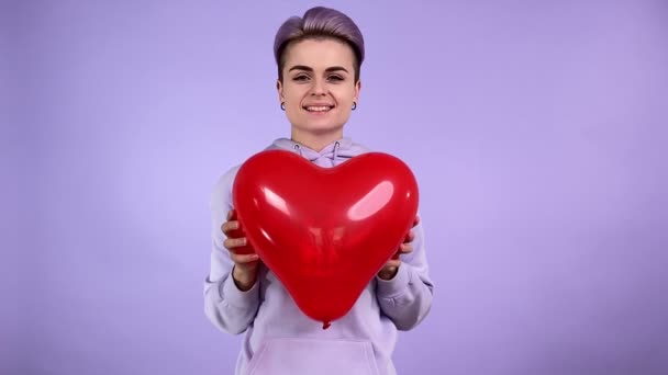 年轻快乐的女同性恋者 紫色短发 心脏形状呈红色气球 手拉手靠近摄像机 心情愉快 在室内的单色紫色背景下面带微笑 — 图库视频影像