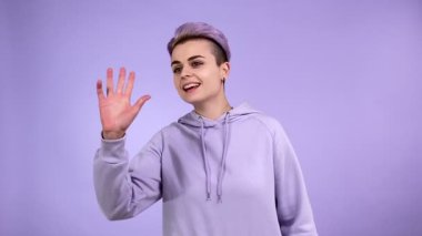 Renkli kısa saçlı, mor eşofman altı giyen, el kol hareketi yapan, buraya gelen, mutlu bir şekilde davet eden, kapalı alanda tek renkli arka planda gülümseyen şık bir genç kadın.