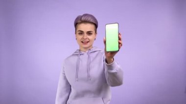 Havalı, genç bir kadın zoomlayıcı moda görünümlü, krom tuşlu akıllı telefonu gösteren, yeşil ekrana işaret eden, kapalı alanda mor arka planda izole edilmiş en iyi fiyat öneren