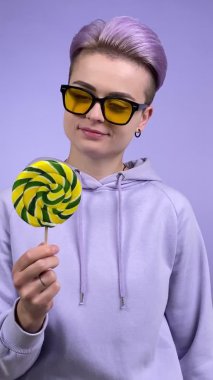 Modern yetişkin kadın geninin dikey görüntüsü mor kumaş içinde, kısa menekşe saçlı, sarı gözlüklü, lolipop tutan, şeker çeviren, kameraya bakan, tek renkli arka planda yalayan