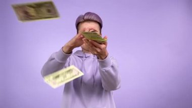 Neşeli genç yetişkin kadın nesli Z, modaya uygun kısa saçlı, parayı çöpe atan, şans piyangosuyla sevinen, para kazanmakla övünen. Elinde bir banknot var. Mor arka planda izole edilmiş kapalı stüdyo çekimi